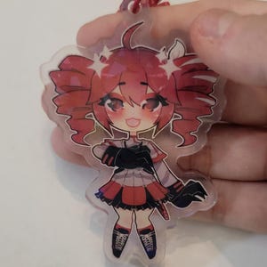 May include: A clear acrylic charm featuring a cartoon character with red hair styled in pigtails. The character wears a black and red outfit with matching shoes. A red string is attached for hanging.