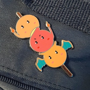 May include: A gold-colored enamel pin featuring three cartoon dango (Japanese dumplings) on a stick. The top dango is yellow, the middle dango is red, and the bottom dango is yellow. The dango have cute faces and the bottom dango has small blue wings. The pin is signed "AlexiAnne".