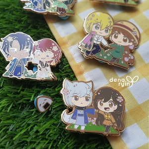 May include: A collection of enamel pins with anime-style character designs, each with a gold-coloured metal outline. The characters are shown in pairs, set on a grassy base. One pin features a rice ball. The background includes a yellow and white checked pattern and green grass.