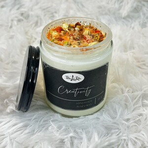 May include: A white candle in a glass jar with a black label that says "Creativity" and "Handmade in Nevada". The candle is topped with dried flowers and herbs.