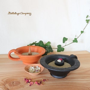 Könnte beinhalten: Zwei keramische Räuchergefäße, eines orange und eines schwarz, mit Sand und Räucherkegeln. Das orangefarbene Gefäß hat einen Griff und das schwarze Gefäß einen kleinen Griff. Neben den Gefäßen befindet sich eine kleine Schale mit weißem Harz.