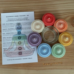 Op de afbeelding: Set van zeven theelichtjes in een regenboog van kleuren, met een witte kaart die de chakra-kleuren en hun betekenis beschrijft. De theelichtjes zijn in een cirkel gerangschikt, met een glazen theelichthouder in het midden.