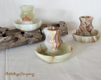 May include: Three decorative candle holders made of green and brown onyx. Each holder has a small, round dish and a taller, cylindrical cup. The holders are arranged on a piece of driftwood.