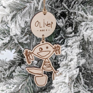 May include: A wooden Christmas ornament with a child's drawing of a person on a bike. The ornament also has a round tag with the name "Oliver - Age 5" written on it.