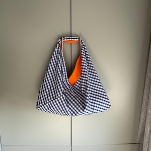 May include: A blue and white gingham tote bag with an orange lining hangs from a silver metal handle. The bag is triangular and made of fabric. The background is a light grey wall with a cabinet.