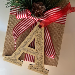 May include: A wooden letter "A" ornament with floral detailing hangs in front of a burlap-wrapped gift box. The box is decorated with a red and white striped ribbon and a sprig of evergreen. A pine cone sits atop the box.