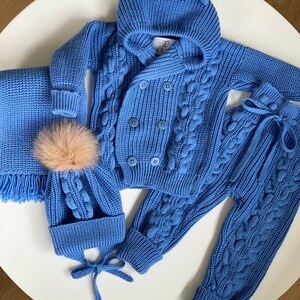 K&ouml;nnte beinhalten: Hellblaues Strick-Outfit-Set f&uuml;r Babys, bestehend aus einer Kapuzenjacke mit B&auml;renohren, einer passenden M&uuml;tze mit Bommel, einem Schal und einer Hose. Das Set hat ein Zopfmuster und Knopfverschl&uuml;sse.