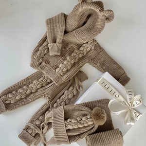 K&ouml;nnte beinhalten: Ein beiges Strick-Baby-Outfit-Set, bestehend aus einer Kapuzenjacke mit B&auml;renohren, einer Hose, einer M&uuml;tze mit Bommel und einem Schal. Jacke und Hose haben dekorative Bommel. Das Set wird auf einer wei&szlig;en Oberfl&auml;che mit einer Geschenkbox pr&auml;sentiert.