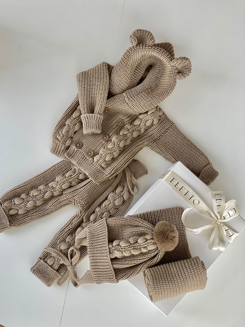 K&ouml;nnte beinhalten: Ein beiges Strick-Baby-Outfit-Set, bestehend aus einer Kapuzenjacke mit B&auml;renohren, einer Hose, einer M&uuml;tze mit Bommel und einem Schal. Jacke und Hose haben dekorative Bommel. Das Set wird auf einer wei&szlig;en Oberfl&auml;che mit einer Geschenkbox pr&auml;sentiert.