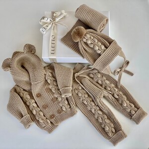 K&ouml;nnte beinhalten: Beige gestricktes Baby-Kleidungsset, bestehend aus einer Kapuzenjacke mit B&auml;renohren, einer Hose, einer M&uuml;tze mit Bommel und einem Schal. Das Set wird mit einer Geschenkbox mit Schleife pr&auml;sentiert.