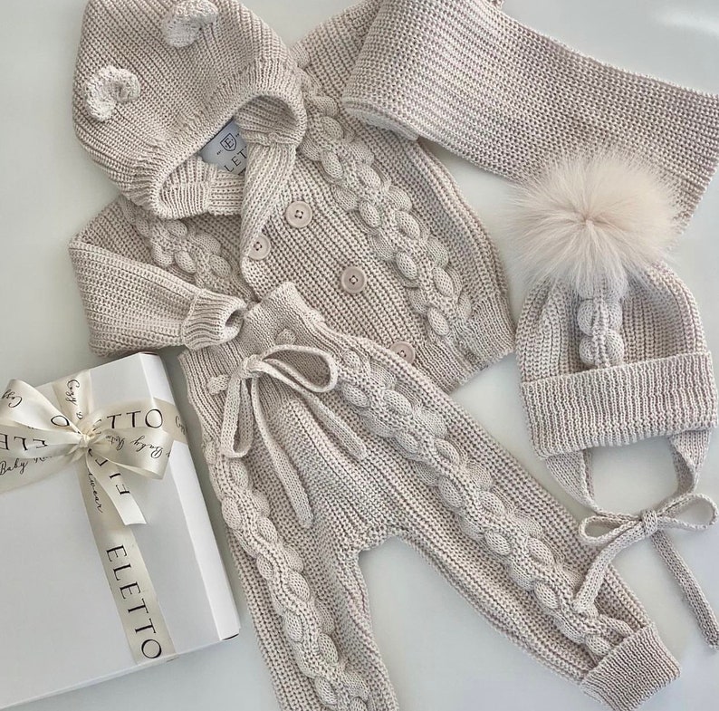 K&ouml;nnte beinhalten: Beiges Strick-Baby-Outfit, bestehend aus einer Kapuzenjacke, einer Hose, einem Schal und einer M&uuml;tze mit einem Kunstfell-Bommel. Die Jacke hat Knopfverschl&uuml;sse und die Hose einen Kordelzug. Das Set wird mit einer Geschenkbox pr&auml;sentiert.