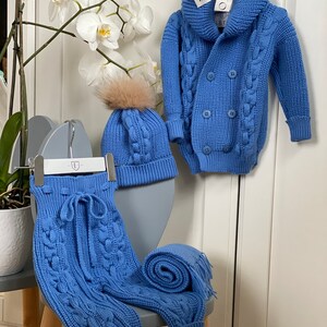 K&ouml;nnte beinhalten: Ein babyblaues Strick-Set f&uuml;r Babys, bestehend aus einer Kapuzenjacke mit doppelreihiger Knopfleiste, einer M&uuml;tze mit Kunstfell-Bommel und einer Hose mit Kordelzug. Ein passender Schal ist zusammengerollt. Das Set wird auf einem grauen Stuhl pr&auml;sentiert.