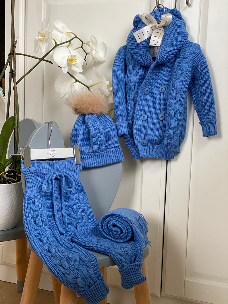 K&ouml;nnte beinhalten: Ein babyblaues Strick-Set f&uuml;r Babys, bestehend aus einer Kapuzenjacke mit doppelreihiger Knopfleiste, einer M&uuml;tze mit Kunstfell-Bommel und einer Hose mit Kordelzug. Ein passender Schal ist zusammengerollt. Das Set wird auf einem grauen Stuhl pr&auml;sentiert.