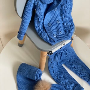 K&ouml;nnte beinhalten: Ein blaues Strick-Baby-Outfit, bestehend aus einem doppelreihigen Cardigan, einer Hose, einem Schal und einer M&uuml;tze mit einem Kunstfell-Bommel. Das Set wird auf einem wei&szlig;en Tisch und einem grauen Stuhl pr&auml;sentiert.