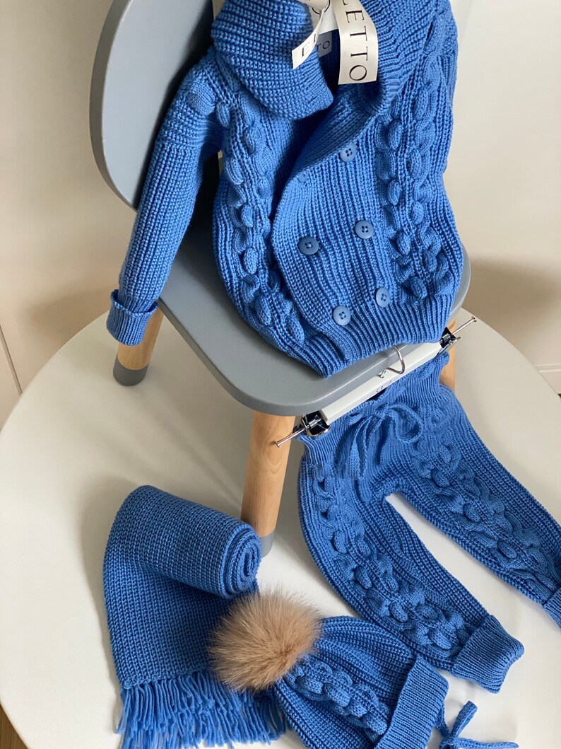 K&ouml;nnte beinhalten: Ein blaues Strick-Baby-Outfit, bestehend aus einem doppelreihigen Cardigan, einer Hose, einem Schal und einer M&uuml;tze mit einem Kunstfell-Bommel. Das Set wird auf einem wei&szlig;en Tisch und einem grauen Stuhl pr&auml;sentiert.