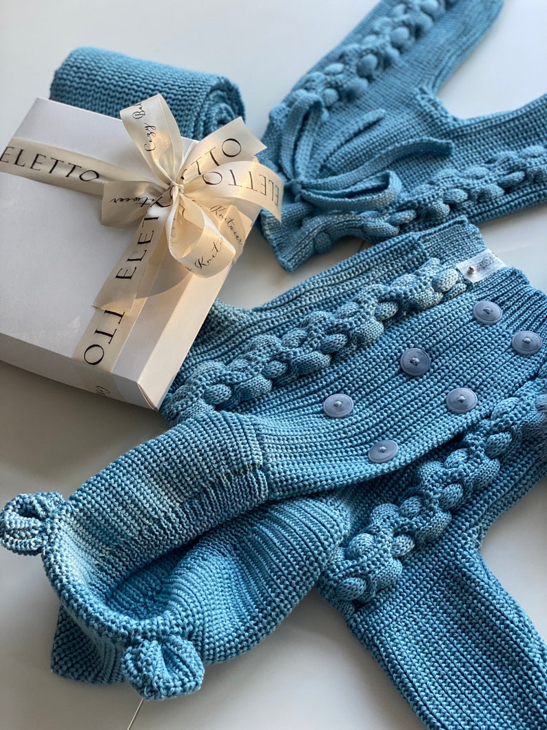 K&ouml;nnte beinhalten: Hellblaues Strick-Baby-Outfit, bestehend aus einer Strickjacke mit Kn&ouml;pfen und dekorativen Bommeln, einer Hose und einer gerollten Decke. Eine wei&szlig;e Geschenkbox mit Schleife ist ebenfalls vorhanden.