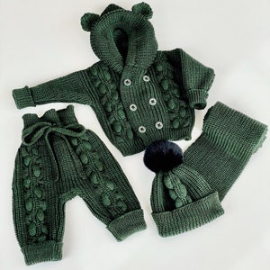 K&ouml;nnte beinhalten: Dunkelgr&uuml;nes Strick-Babyset, bestehend aus einer Kapuzenjacke mit B&auml;renohren, einer Hose, einer M&uuml;tze mit schwarzem Bommel und einem Schal. Das Set hat ein Zopfmuster und ist aus weichem Garn gefertigt.