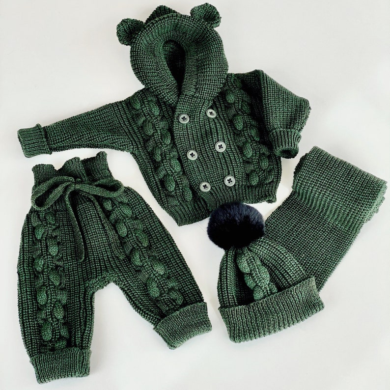 K&ouml;nnte beinhalten: Dunkelgr&uuml;nes Strick-Babyset, bestehend aus einer Kapuzenjacke mit B&auml;renohren, einer Hose, einer M&uuml;tze mit schwarzem Bommel und einem Schal. Das Set hat ein Zopfmuster und ist aus weichem Garn gefertigt.