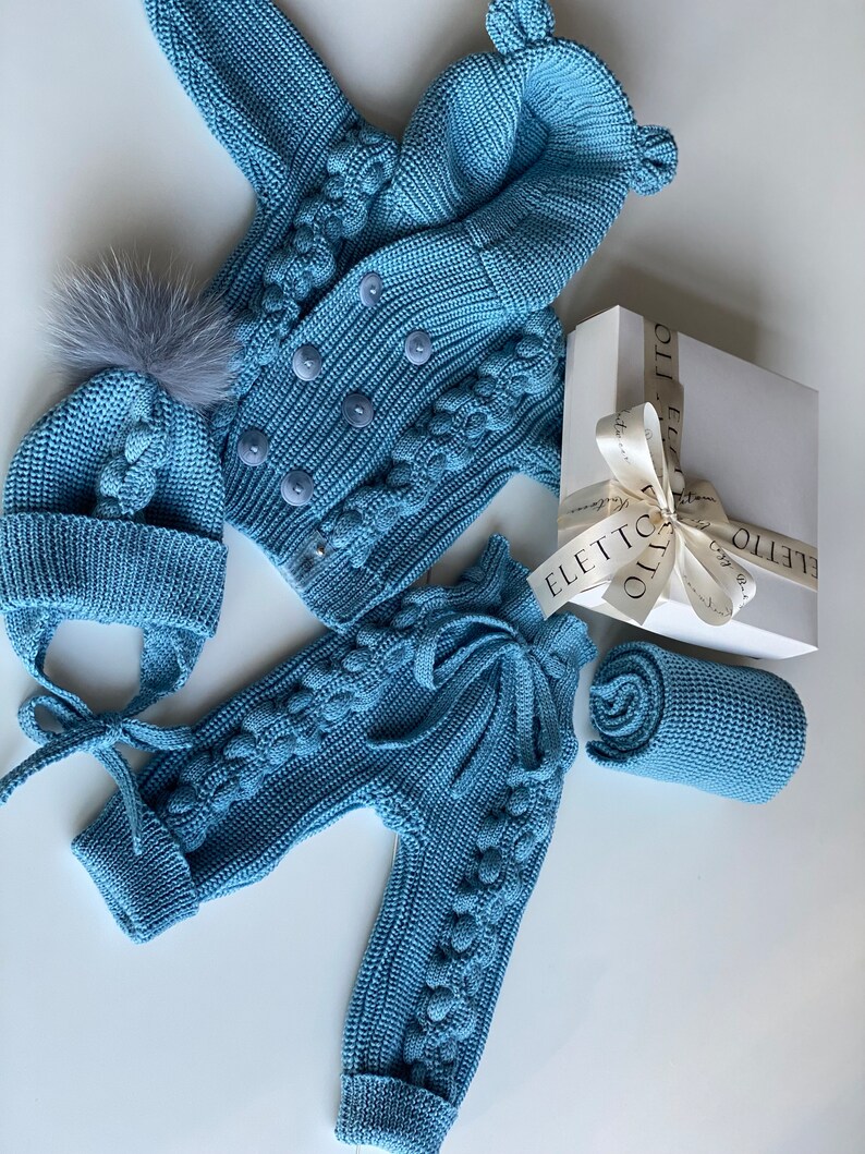 K&ouml;nnte beinhalten: Hellblaues Strick-Outfit-Set f&uuml;r Babys, bestehend aus einer Jacke mit Kn&ouml;pfen, einer Hose mit Bindeb&auml;ndern, einer M&uuml;tze mit Bommel und einer gerollten Decke. Das Set wird mit einer Geschenkbox pr&auml;sentiert.