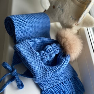 K&ouml;nnte beinhalten: Ein blaues Strickset, bestehend aus einer M&uuml;tze mit Kunstfell-Bommel und einem passenden Schal. Der Schal ist gerollt und hat Fransen an den Enden. Die M&uuml;tze hat Ohrenklappen und Bindeb&auml;nder. Das Set ist aus weichem, strukturiertem Garn.