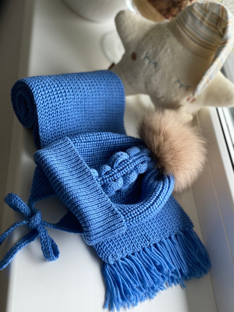 K&ouml;nnte beinhalten: Ein blaues Strickset, bestehend aus einer M&uuml;tze mit Kunstfell-Bommel und einem passenden Schal. Der Schal ist gerollt und hat Fransen an den Enden. Die M&uuml;tze hat Ohrenklappen und Bindeb&auml;nder. Das Set ist aus weichem, strukturiertem Garn.