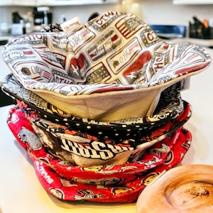 May include: A stack of four fabric bowl cozies. The top cozy is white with red, blue, and gray patterned fabric featuring the word "Ohio". The second cozy is black with white polka dots. The third cozy is black with white and red patterned fabric featuring the word "The" and a football. The bottom cozy is red with white and black patterned fabric featuring the word "Buckeye".