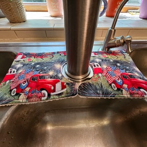 May include: A patriotic sink splash guard featuring a red truck with American flags, fireworks, and a picnic basket. The design is set against a dark green background. The guard is designed to fit around a faucet.