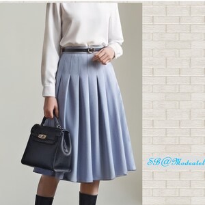 Peut inclure: Jupe pliss&eacute;e bleu clair, associ&eacute;e &agrave; un haut blanc &agrave; manches longues et une ceinture noire. Un sac &agrave; main noir est tenu. La jupe arrive aux genoux. Le fond est un mur de briques blanches.