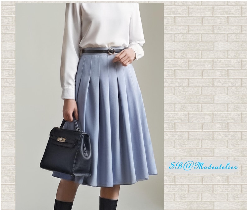 Peut inclure: Jupe pliss&eacute;e bleu clair, associ&eacute;e &agrave; un haut blanc &agrave; manches longues et une ceinture noire. Un sac &agrave; main noir est tenu. La jupe arrive aux genoux. Le fond est un mur de briques blanches.