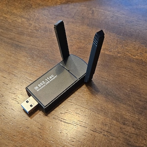 May include: A black 802.11ac wireless USB adapter with two adjustable antennas. The adapter has a USB connector and is resting on a wooden surface. The text "802.11ac Wireless USB Adapter" is printed on the device.