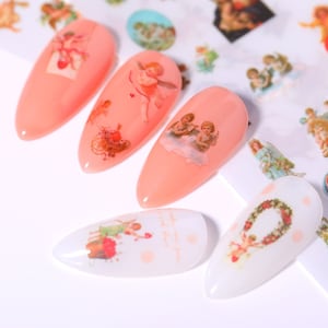 May include: A set of six pink and white nail decals featuring various vintage cherub designs. The decals include a cherub with a bow and arrow, a cherub holding a heart, a cherub with a wreath, and a cherub with a flower.