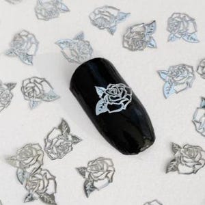 May include: Silver metallic rose nail art decals. The decals are in the shape of roses with leaves. The decals are scattered on a white surface with one decal on a black nail.