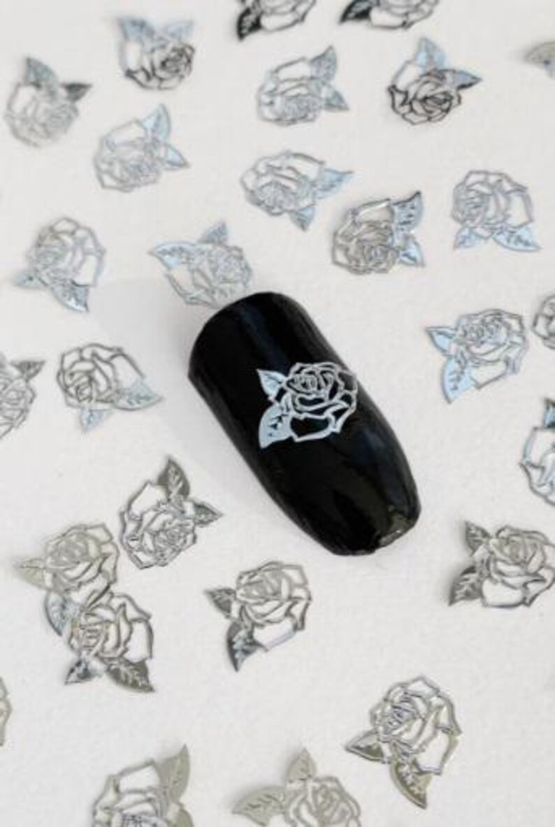 May include: Silver metallic rose nail art decals. The decals are in the shape of roses with leaves. The decals are scattered on a white surface with one decal on a black nail.