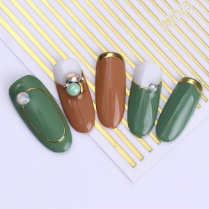 May include: Five different nail designs with gold accents. The nails are painted in shades of green and brown. Some nails have small, clear rhinestones.