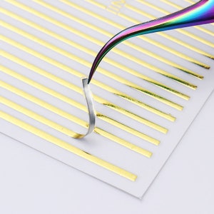 May include: A sheet of gold nail art strips with a rainbow-colored tweezers picking up one of the strips.