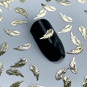 May include: A scattering of gold angel wing nail charms on a light gray surface. A black nail with a gold angel wing charm is in the center of the image.