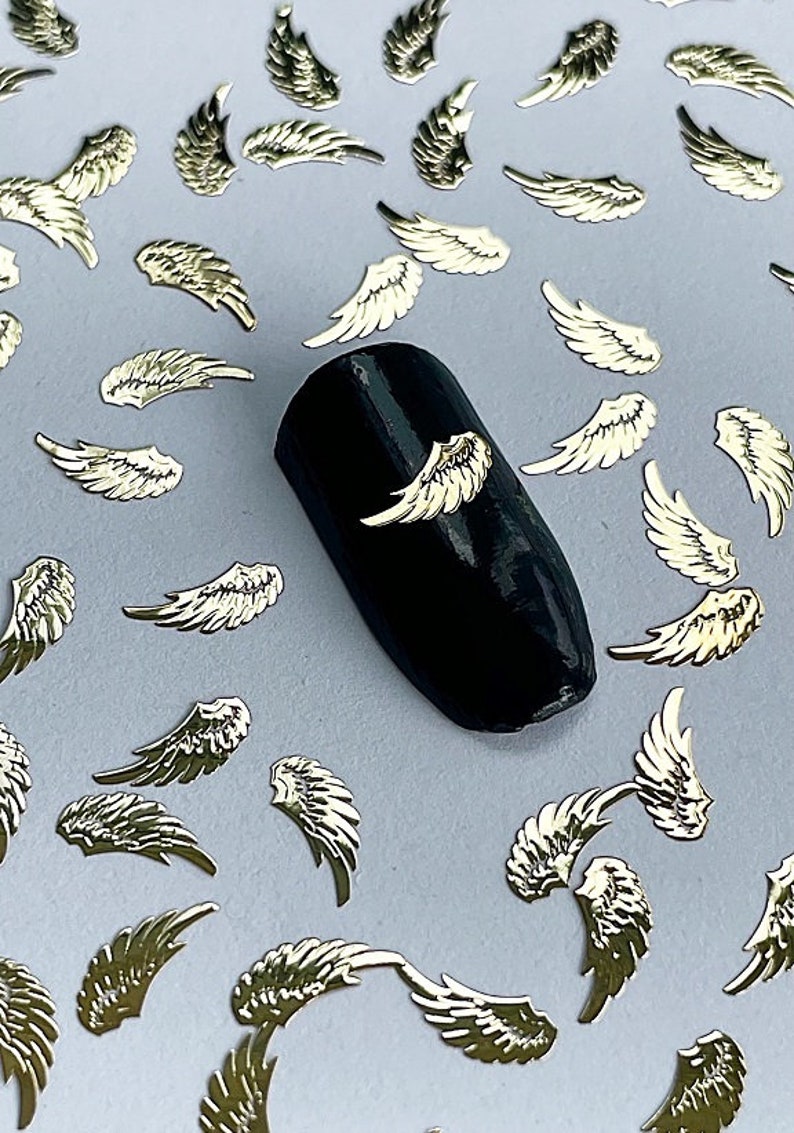 May include: A scattering of gold angel wing nail charms on a light gray surface. A black nail with a gold angel wing charm is in the center of the image.