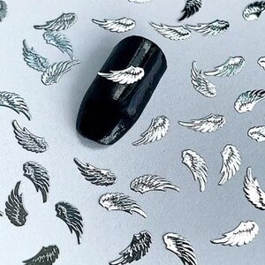May include: Silver angel wings nail art embellishments scattered on a light gray surface. A black nail with a silver angel wing is shown in the center of the image.