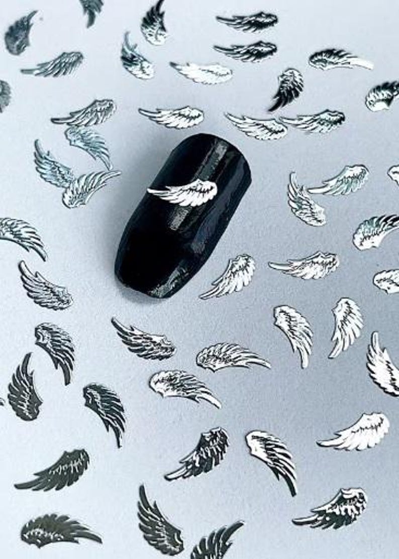 May include: Silver angel wings nail art embellishments scattered on a light gray surface. A black nail with a silver angel wing is shown in the center of the image.