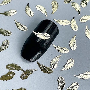May include: Gold angel wing nail art embellishments scattered on a light gray surface. A black nail with a gold angel wing on it is in the center of the image.