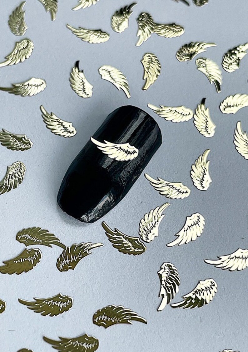 May include: Gold angel wing nail art embellishments scattered on a light gray surface. A black nail with a gold angel wing on it is in the center of the image.