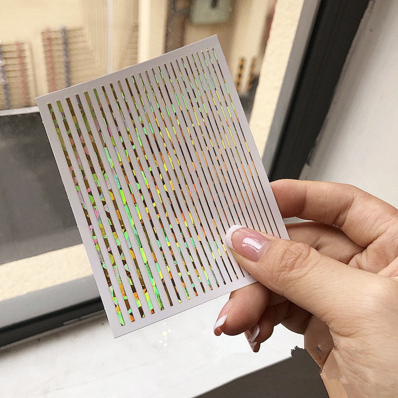 May include: A sheet of iridescent nail art stickers with thin, vertical lines. The lines are a rainbow of colors, including green, yellow, orange, and pink.