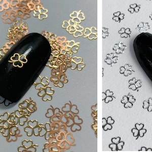 May include: Two images showing nail art with gold and silver four-leaf clover charms. The charms are scattered around a black nail polish on a white background.