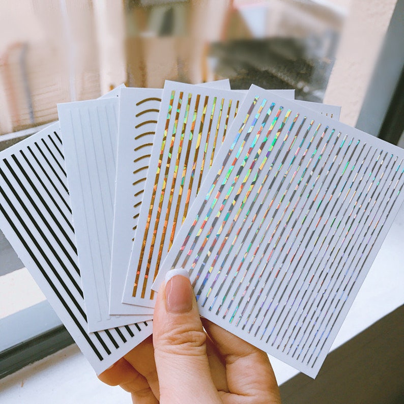 May include: Set of six sheets of nail art stickers with different designs. The stickers feature black, gold, and iridescent lines on a white background.