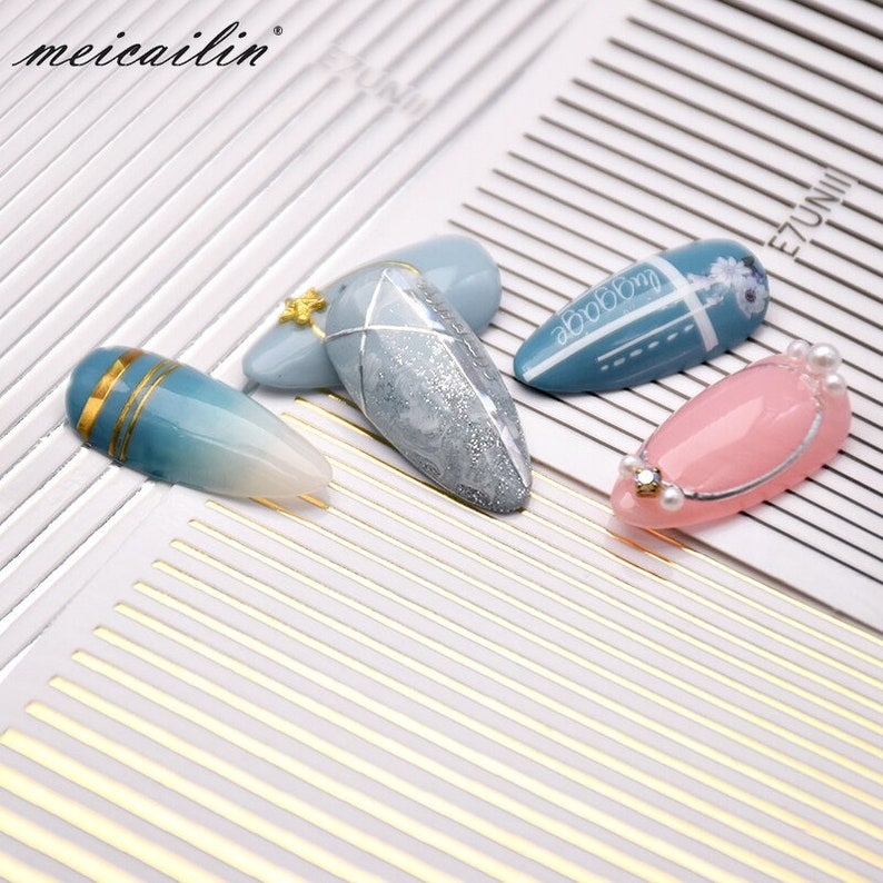 May include: Four different nail art designs on a white background with gold lines. The designs include a blue ombre with gold stripes, a light blue design with a silver glitter finish, a light blue design with a white and black geometric pattern, and a pink design with a silver line and pearl accents.