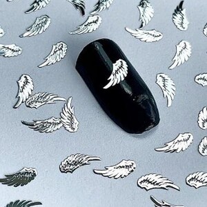 May include: Silver angel wings nail art embellishments. The wings are scattered on a white surface and a black nail with a silver angel wing is shown in the center of the image.