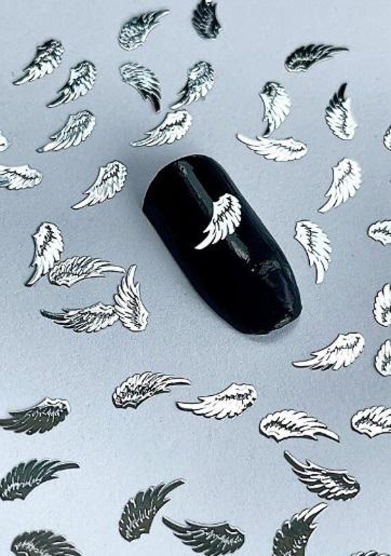 May include: Silver angel wings nail art embellishments. The wings are scattered on a white surface and a black nail with a silver angel wing is shown in the center of the image.