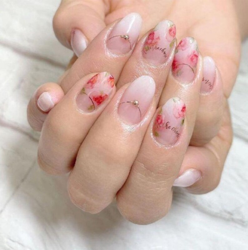 May include: Close-up of a hand with pink and white oval-shaped nails. Each nail is decorated with a floral design and a small silver bead.