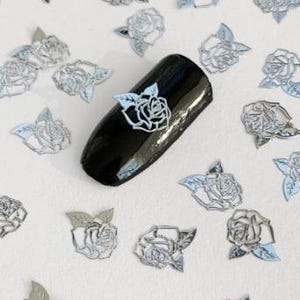 May include: Silver metallic rose nail art decals. The decals are in the shape of a rose with leaves. The decals are scattered on a white surface. One decal is placed on a black nail.