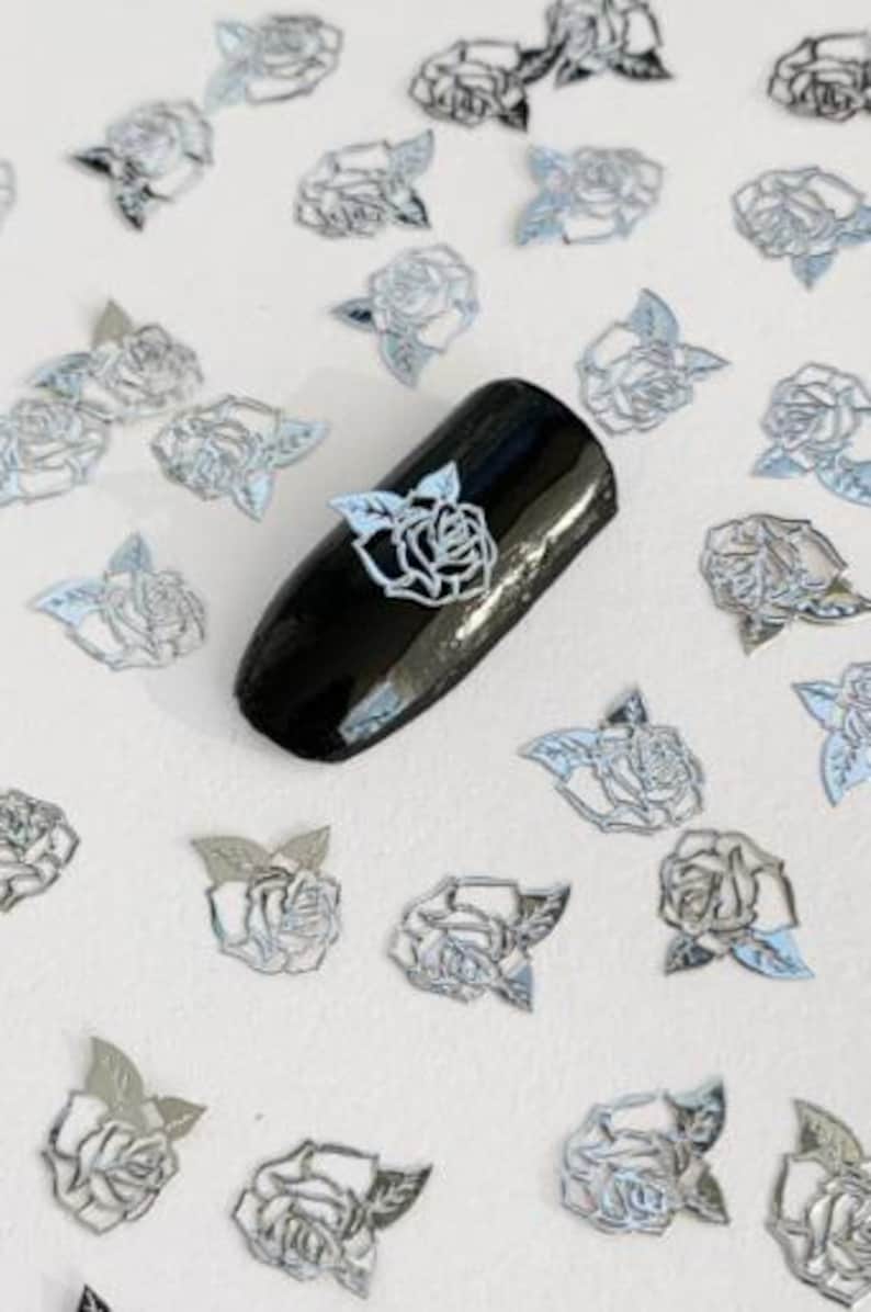 May include: Silver metallic rose nail art decals. The decals are in the shape of a rose with leaves. The decals are scattered on a white surface. One decal is placed on a black nail.